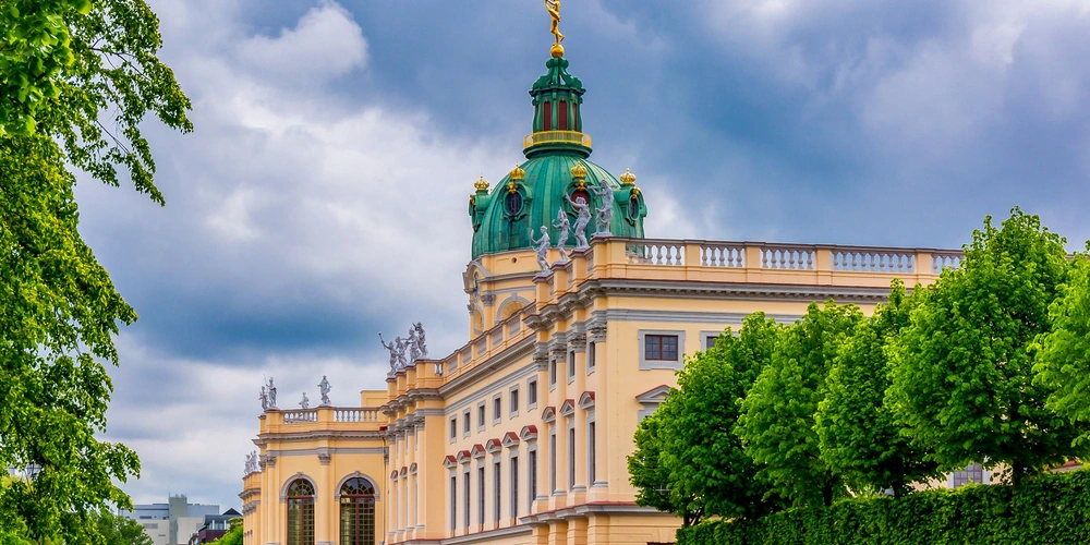 Historisches Gebäude mit grünem Dach und umliegenden Bäumen unter bewölktem Himmel.