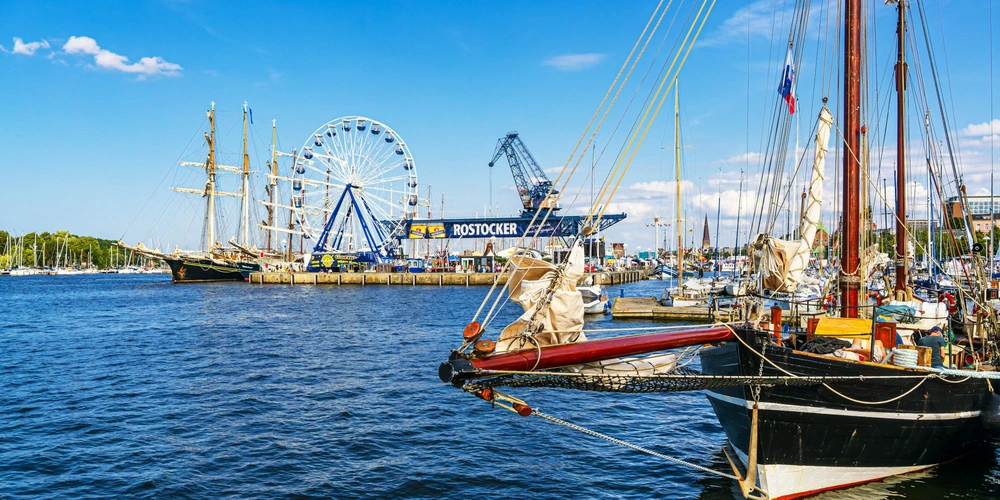 Hafenansicht mit Segelschiffen, Riesenrad und Industriekran unter blauem Himmel.