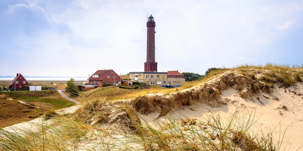 Leuchtturm hinter Dünen und kleinen Gebäuden an der Küste, unter blauem Himmel.