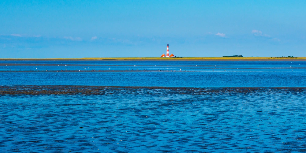 Schleswig-Holsteinisches Wattenmeer und Westerhever Leuchtturm an der Küste mit blauem Wasser und klarem Himmel im Hintergrund.