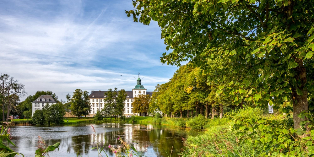Schloss mit Wasserfläche und Bäumen unter einem bewölkten Himmel.