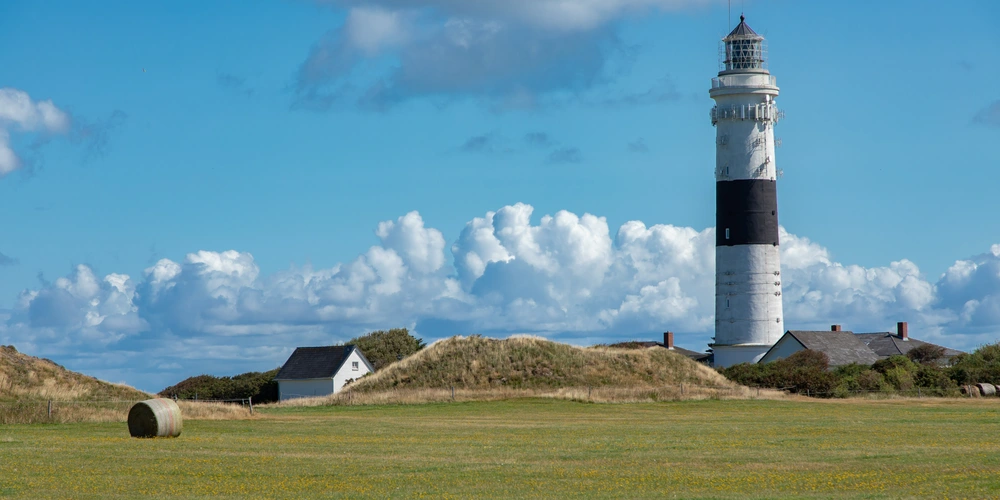 Leuchtturm vor blauem Himmel mit Wolken, umgeben von grünem Feld und Dünen.
