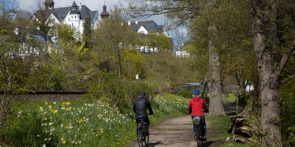 Plön mit Blick auf das Plöner Schloss Radfahrer auf einem Weg, umgeben von Bäumen und Blumen, mit einem Schloss im Hintergrund.