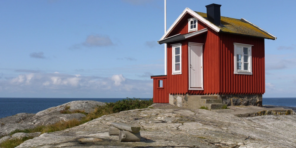 Rote Holzhütte auf einem Felsen mit Meerblick und klarem Himmel.