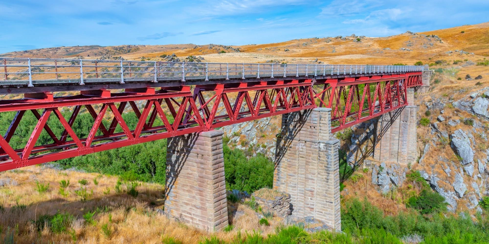 Rote Eisenbrücke über eine Schlucht, umgeben von hügeliger Landschaft und blauem Himmel.
