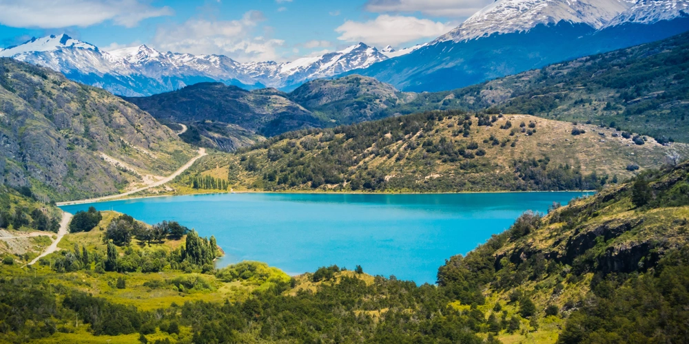 Türkisblaues Wasser eines Sees umgeben von grünen Hügeln und schneebedeckten Bergen.