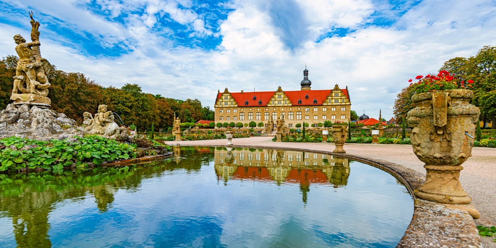 Schloss mit roten Dächern spiegelt sich im klaren Wasser eines Gartens.