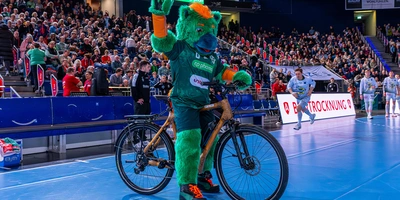 Das Maskottchen der Recken winkt auf einem Fahrrad vor einer jubelnden Menge in der Halle.
