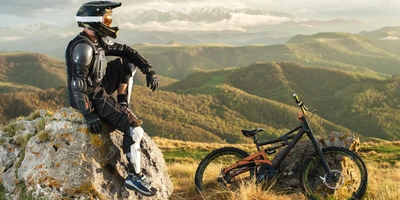 Ein Downhill-Fahrer sitzt auf in einer Downhill-Ausrüstung auf einem Stein neben seinem Mountainbike.