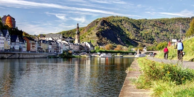 2 Radfahrer fahren entlang eines Flusses, auf dem ein Schiff an einer Stadt liegt.