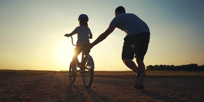 Erkenne, ob dein Kind bereit ist mit linexo Ein Erwachsener hilft einem Kind beim Fahrradfahren, mit Sonnenuntergang im Hintergrund.