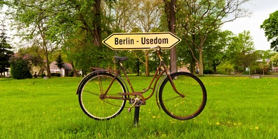 Ein rostiges Fahrrad steht neben einem Wegweiser nach Berlin und Ueckermünde in einer grünen Wiese.