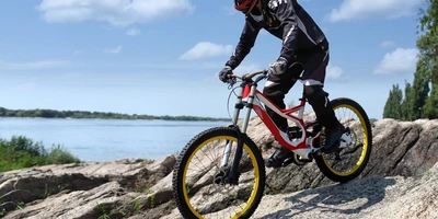 Mountainbiker auf Felsen mit gelben Reifen, im Hintergrund ein Gewässer und blauer Himmel.