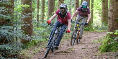 Hier sind 8 nützliche Downhill Pflegetipps. Zwei Personen fahren Mountainbike durch Wald.