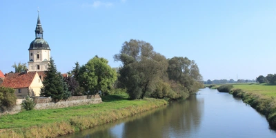 Entdecke die Etappen des Elsterradwegs Ein ruhiger Fluss fließt neben einem historischen Gebäude und Bäumen unter blauem Himmel.