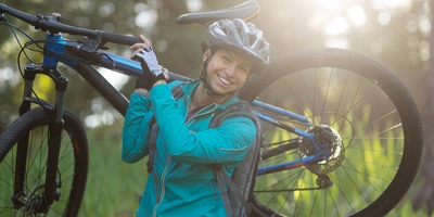 Lächelnde Person trägt ein Mountainbike in einer sonnigen, grünen Landschaft.