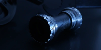 Hollowtech II / GXP / ähnliche Systeme Schwarzes Fahrrad-Bottom-Bracket mit metallischen Details auf dunklem Hintergrund.