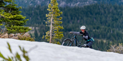 10 Tipps zum E-Bike fahren im Winter. Frau schiebt Mountainbike verschneiten Berg hoch.