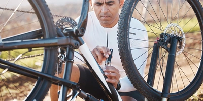 Ein Mensch repariert ein Fahrrad in der Natur, umgeben von Bäumen und Sonne.