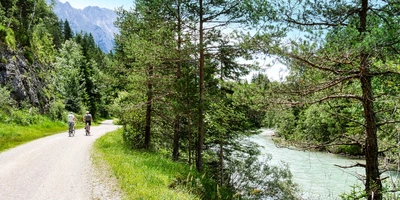 Radfahrer auf einem Schotterweg neben einem klaren Fluss, umgeben von Bäumen und Bergen.