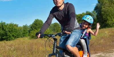 Person fährt ein Kind auf dem Fahrrad, welches im Kindersitz sitzt, durch die Gegend 