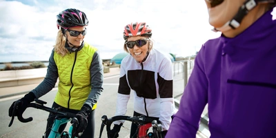 Drei Radfahrerinnen in bunten Sportoutfits lachen auf einer Straße bei bewölktem Himmel