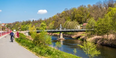 Muldentalradweg  Ein Radfahrer fährt an einem Fluss entlang, mit einer Brücke und grünen Bäumen im Hintergrund.