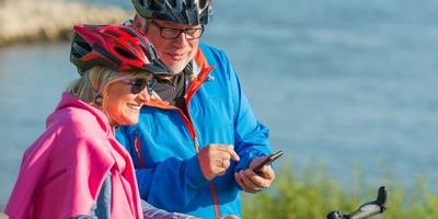 Zwei ältere Menschen am Wasser mit Fahrrädern und Fahrradkleidung schauen auf Handy