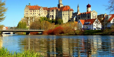 Das Schloss Sigmaringen ist vom Donau-Radweg aus zu sehen. Es ist ein Fluss zu sehen. Im Vordergrund ist das grüne Ufer erkennbar. Auf der anderen Seite des Flusses ist ein Schloss, das Schloss Sigmaringen. Daneben sind kleine Häuser, ein paar Bäume. Eine Brücke führt über den Fluss in Richtung Schloss. Auf dem Fluss, die Donau, spiegelt sich das Schloss.