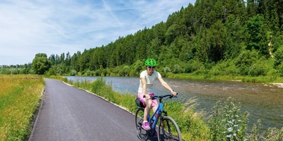 Polenradwege Frau mit Helm radelt neben einem Fluss, umgeben von Bäumen und Wiesen.