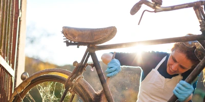 Frau reinigt ein altes Fahrrad im Sonnenlicht mit blauen Handschuhen.