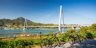 Zwei Fahrräder stehen auf einem Weg, mit einer Brücke und Wasser im Hintergrund.