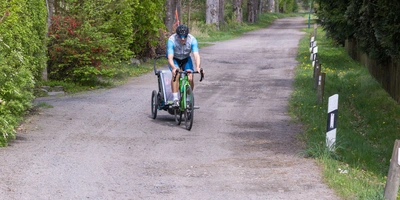 Thule Fahrradanhänger Vergleich: Die besten Modelle Ein Radfahrer mit Anhänger fährt einen schmalen, von Bäumen gesäumten Weg entlang.