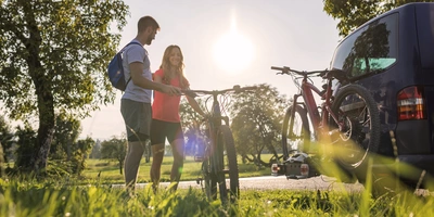 Paar mit Fahrrädern vor einem Auto, umgeben von Bäumen im Sonnenschein.