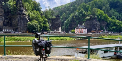 Tschechien-Radwege – die schönsten Routen Fahrrad mit Gepäck nahe einem Fluss, umgeben von grünen Bergen und einem kleinen Ort.