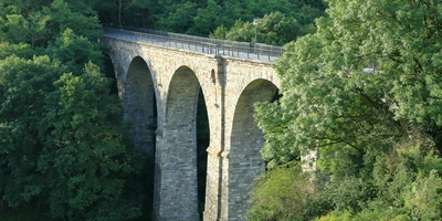 Steinernes Viadukt mit mehreren Bögen, umgeben von Bäumen und Grün.