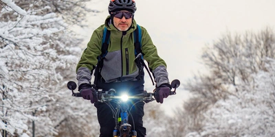 Person auf einem Fahrrad in verschneiter Landschaft, trägt Winterkleidung und Helm.