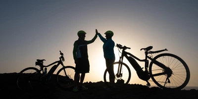 Zwei Personen stehen neben ihren E-Bikes vor dem Sonnenuntergang und klatschen sich ab