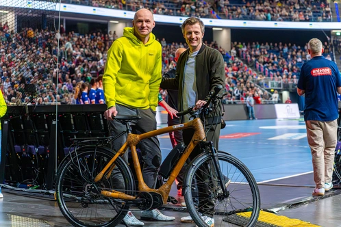 Zwei Männer stehen mit einem Fahrrad in einer großen Sportarena, viele Zuschauer im Hintergrund.