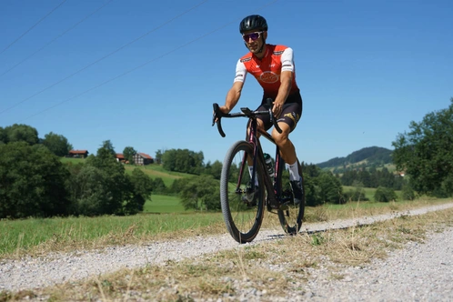 Radfahrer in rot-weißem Trikot fährt über einen Schotterweg in der Natur.