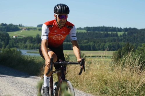 Radfahrer in rot-weißer Kleidung fährt auf einem Schotterweg durch eine Landschaft.